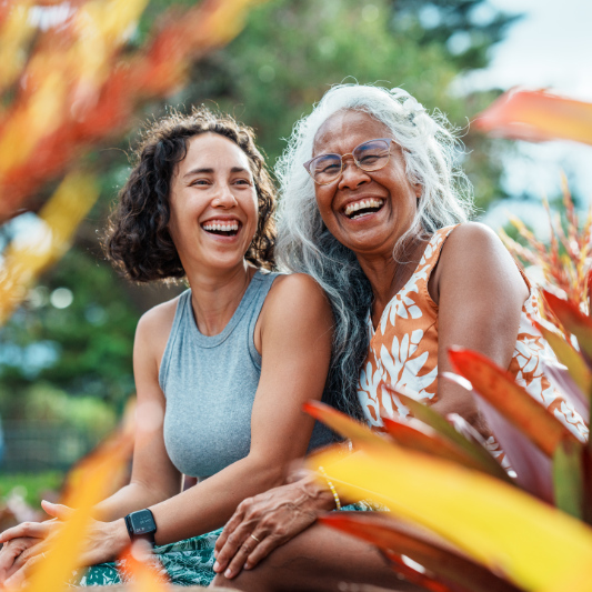 A beautiful and healthy senior woman of Hawaiian and Chinese descent laughs while enjoying quality time outdoors with her Eurasian adult daughter at a park in Hawaii with beautiful tropical landscaping.