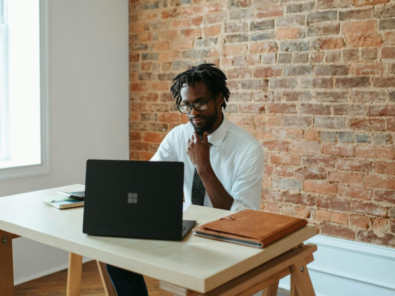 A man sitting at a desk, looking at his laptop with a brick wall behind him